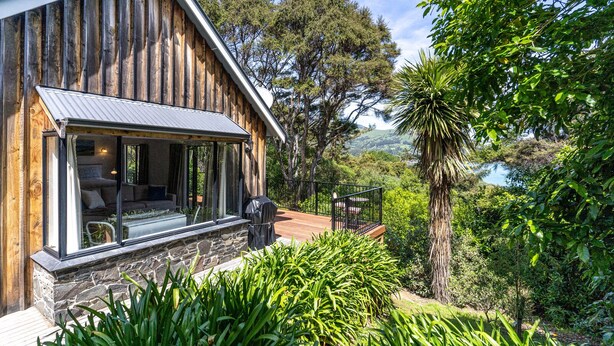 Une cabane en bois, avec des fondations en pierre, de grandes fenêtres et une terrasse offrant une vue sur un jardin luxuriant et des collines.
