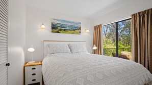 Une chambre à coucher avec un grand lit, une table de chevet, une lampe, un tableau et une vue sur la verdure par la fenêtre.