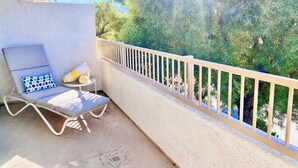 Un balcon avec une balustrade blanche, une chaise longue et une petite table agrémentée d’un coussin et d’une serviette enroulée.