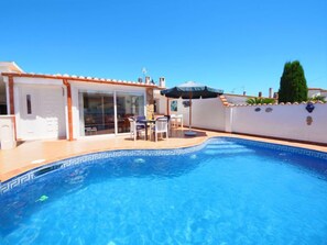 Una piscina con borde de azulejos, un patio con mesa y sillas, y un edificio blanco con techo de tejas.