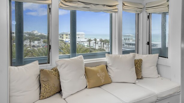 Ein weißes Sofa mit beige und gemusterten Kissen, große Fenster mit Vorhängen und Blick auf einen Strand und Gebäude.