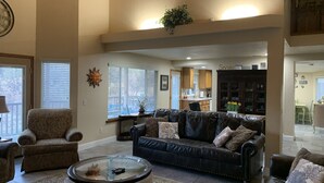 A spacious living room with a dark leather sofa, a glass coffee table, and a patterned armchair. There is a kitchen in the background with wooden cabinets and a dining area. - Peaceful Riverfront Resort #2 (Three Rivers)