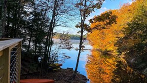 Una terraza de madera con barandilla de metal, un tapete rojo y un contenedor azul sobre una plataforma de madera con vistas a un lago rodeado de árboles de colores otoñales.