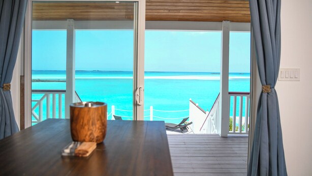 A wooden table with a wooden cup and a napkin, a view of the ocean through a glass door, and a balcony with a white railing. - Ultimate Vacation in Paradise at BLU (Rolletown)