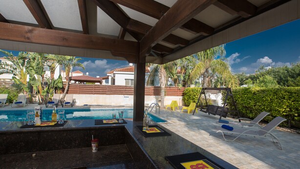 A poolside area with a bar, lounge chairs, and a view of a residential neighborhood. - Ayia Napa Holiday Villa At26 (Sotira)