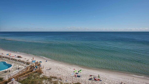 Ein Strand mit klarem, blauem Wasser, einem sandigen Ufer und einem Poolbereich mit Liegestühlen und Sonnenschirmen.