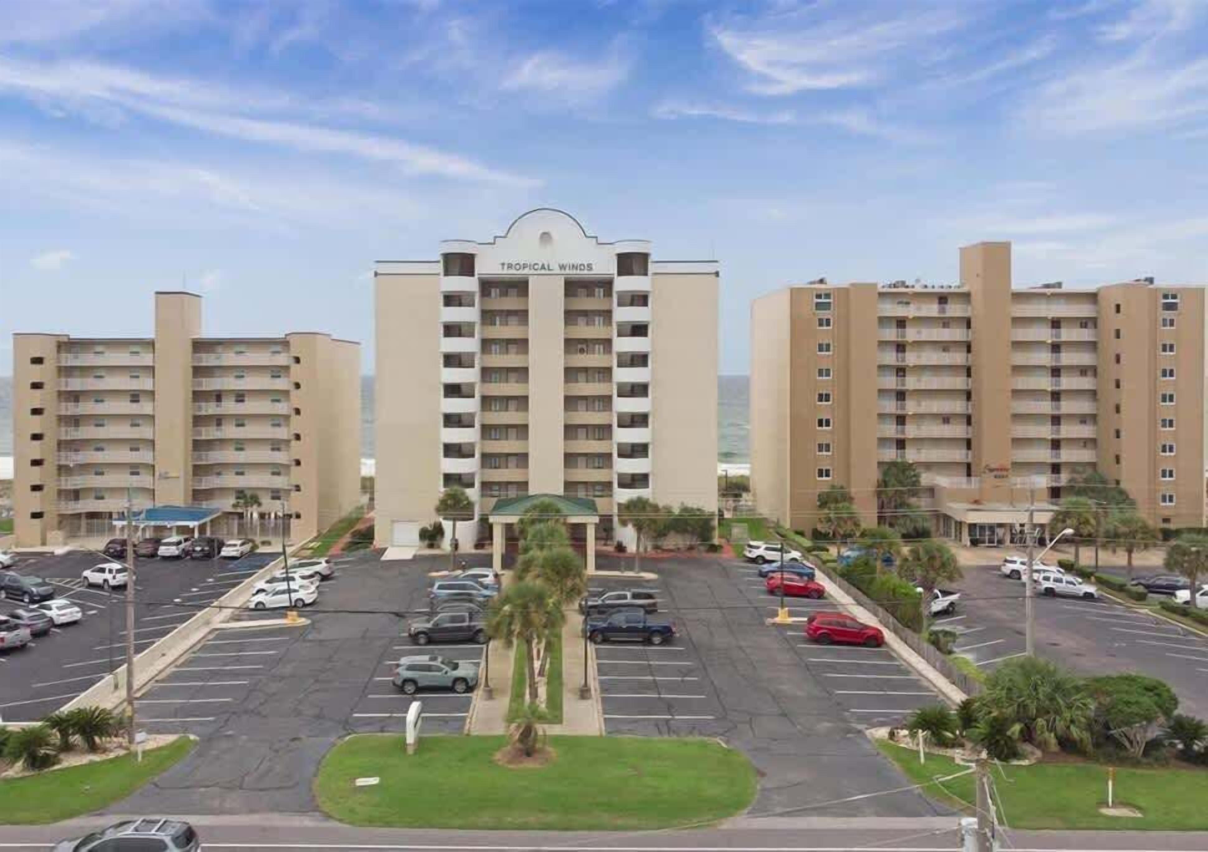 Il s’agit d’un hôtel composé d’un bâtiment principal, portant l’inscription « Tropical Winds », et de plusieurs bâtiments annexes. Un parking avec plusieurs voitures est également disponible.