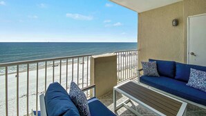 Ein Balkon mit Blick auf den Strand und das Meer, eine Couch mit gemusterten Kissen und eine Tür rechts.
