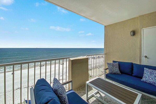 A balcony with a view of the beach and ocean, a sofa with patterned cushions, and a door to the right.