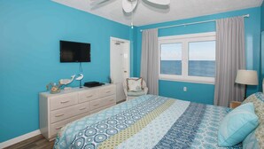 Ein Schlafzimmer mit einem Bett, einem Deckenventilator, einem Fernseher, einem Schminktisch und einem Fenster mit Vorhängen, mit Blick auf das Meer.