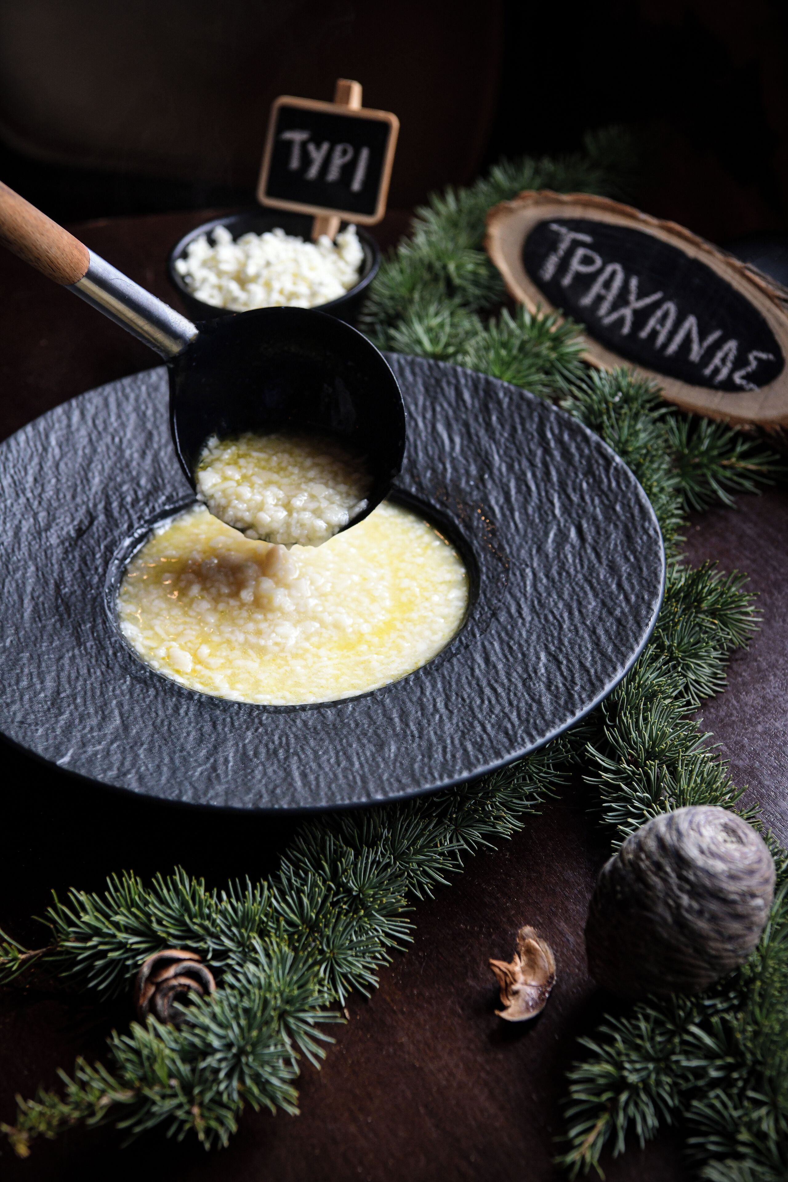 Un tazón de polenta con una cuchara, rodeado de ramas de pino y un letrero de madera que dice 'TYPI'.