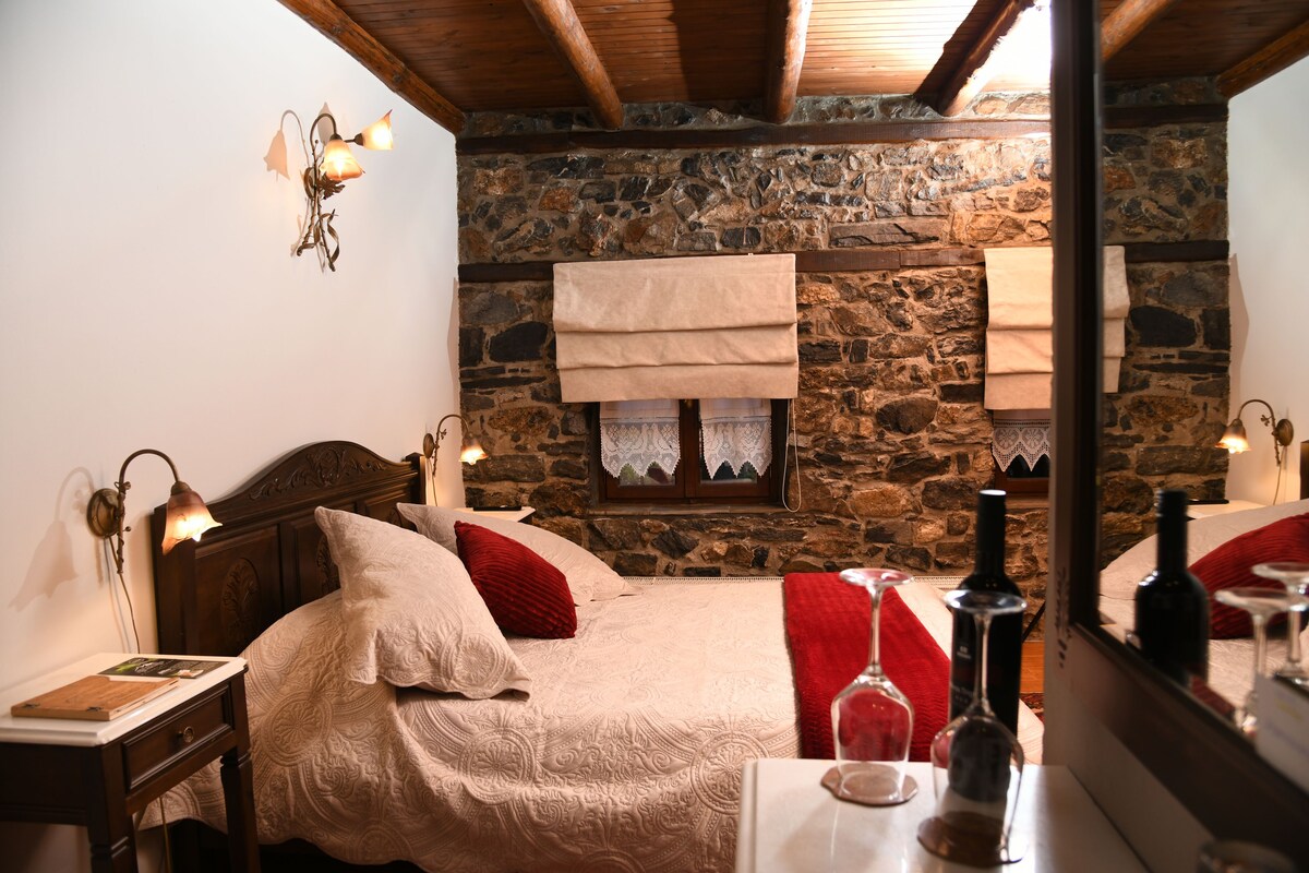 A bedroom with a stone wall, wooden beams, a bed with white and red bedding, a nightstand with a lamp, and a mirror reflecting the room.