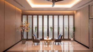 A modern hotel lobby with a polished floor, two leather armchairs, a small round table, a ceiling fan, and a large window with vertical blinds. - Lewit Hotel Pattaya, a member of Radisson Individuals (Pattaya)