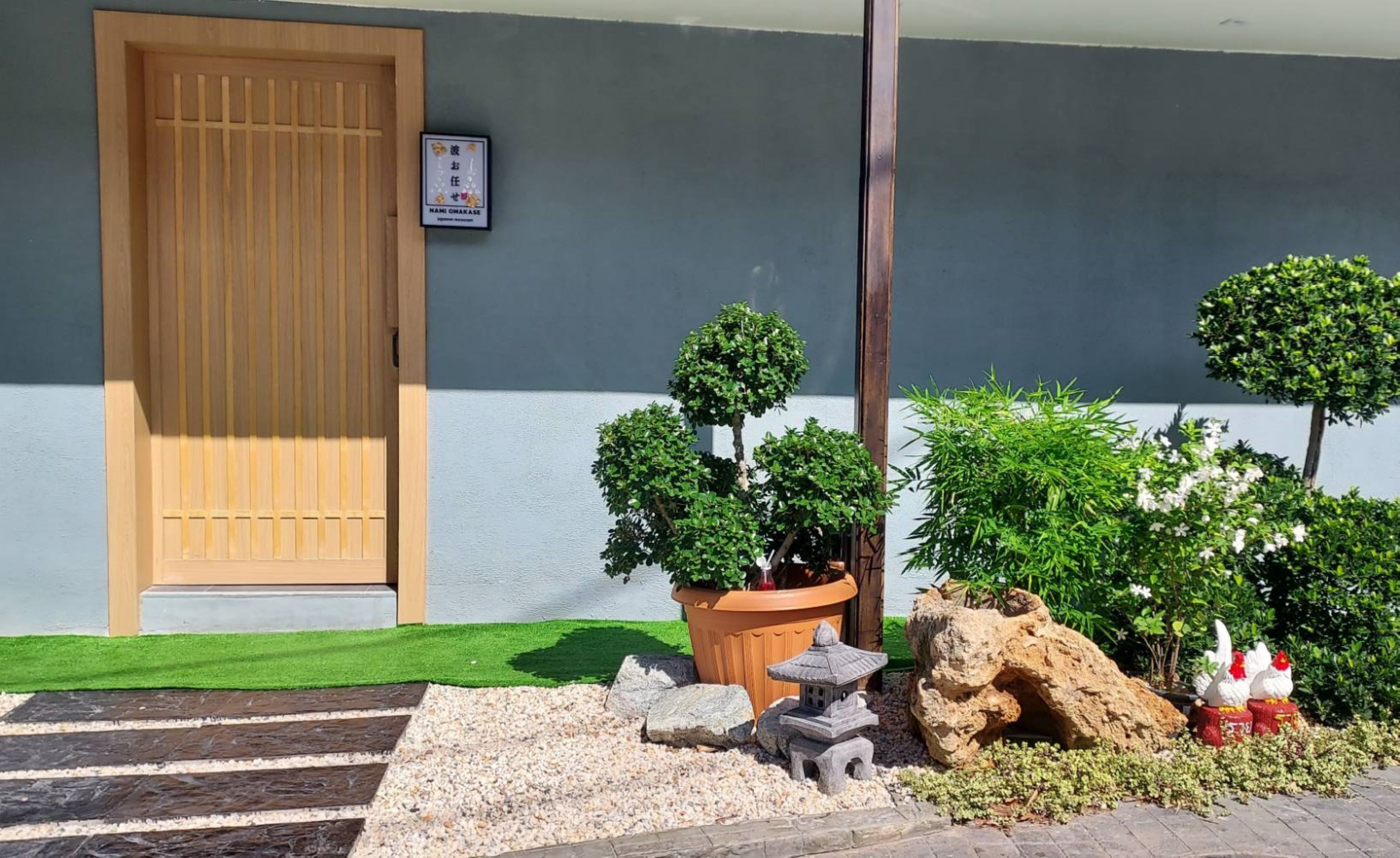 Una entrada tradicional al estilo japonés, con puerta de madera, un pequeño santuario y abundante vegetación cuidada.