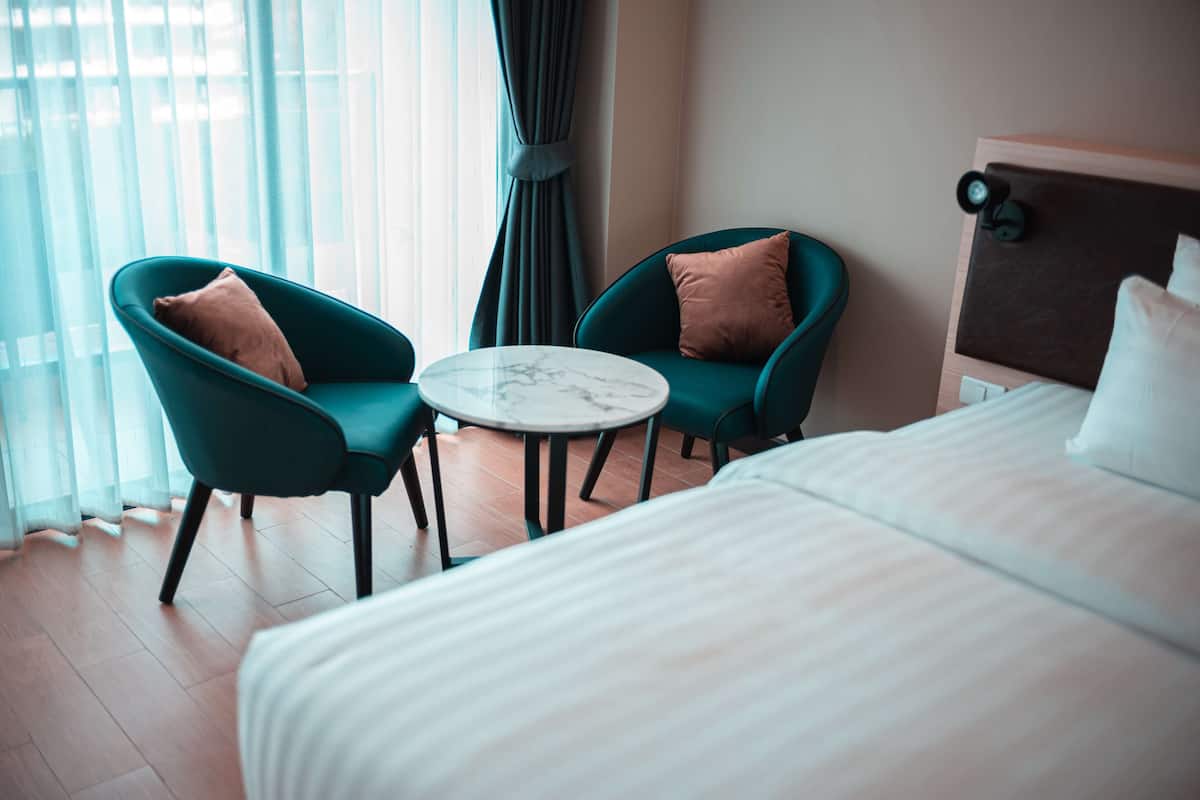 A hotel room with a bed, two teal armchairs, a small round table, and a view of the outside through sheer curtains.