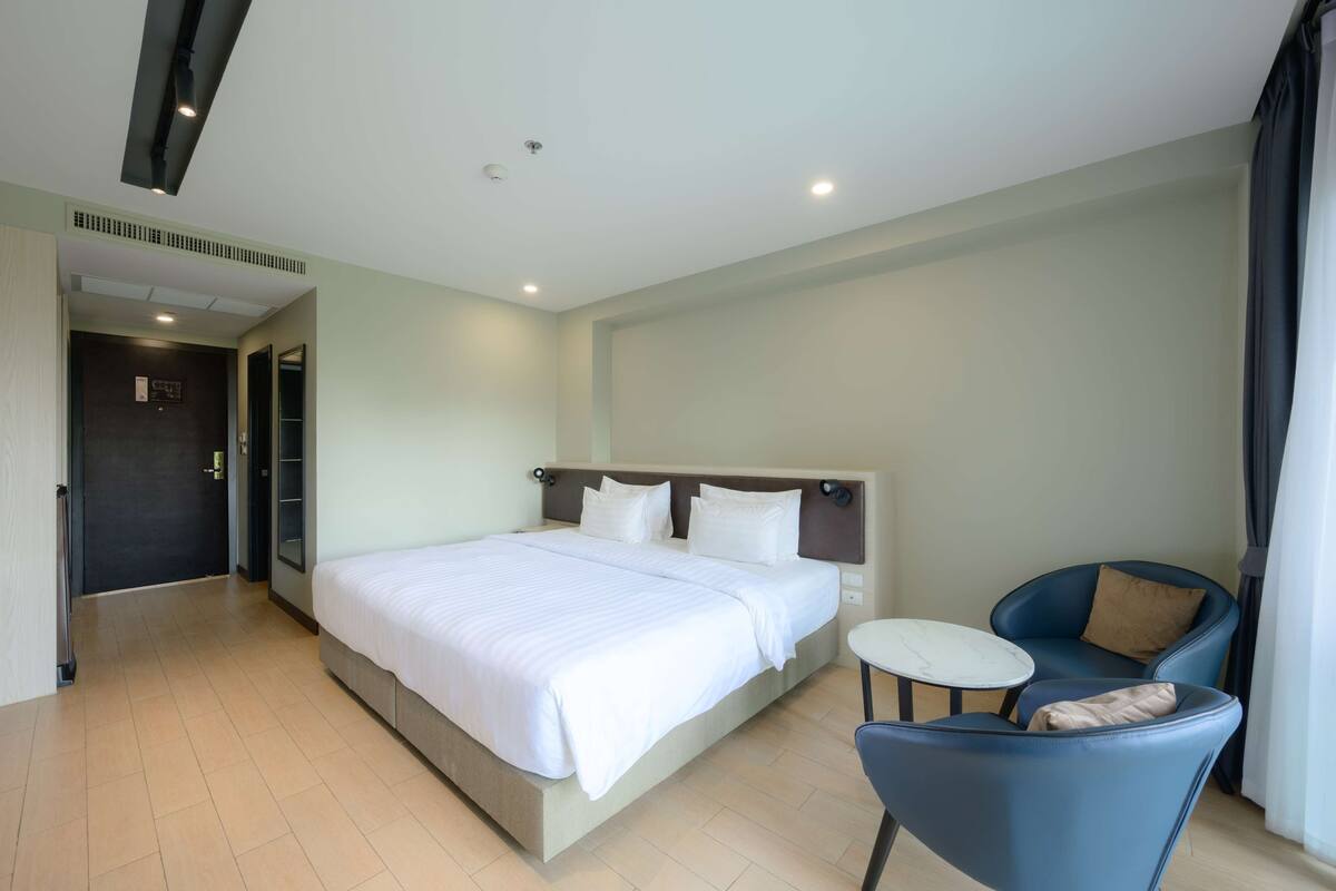 A hotel room with a large bed, a small round table, a blue chair, and a door leading to another room.