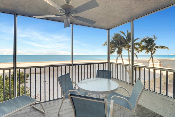 A balcony with a glass table and chairs, overlooking a beach with palm trees and the ocean.