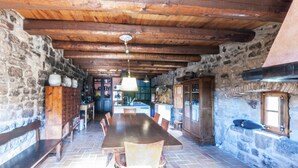 Un coin repas rustique aux murs de pierre, avec une longue table en bois et des bancs, un luminaire suspendu et une cheminée en pierre.