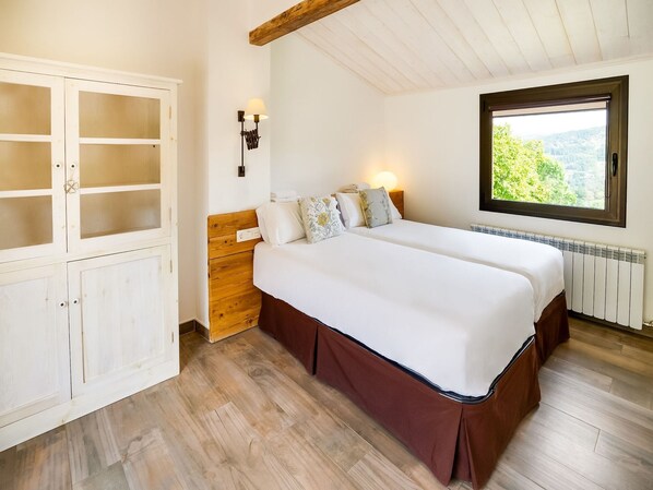 Un dormitorio con cabecera de madera, cama grande con ropa de cama blanca, armario con puertas de cristal, lámpara de pared y ventana con vista a zonas verdes.