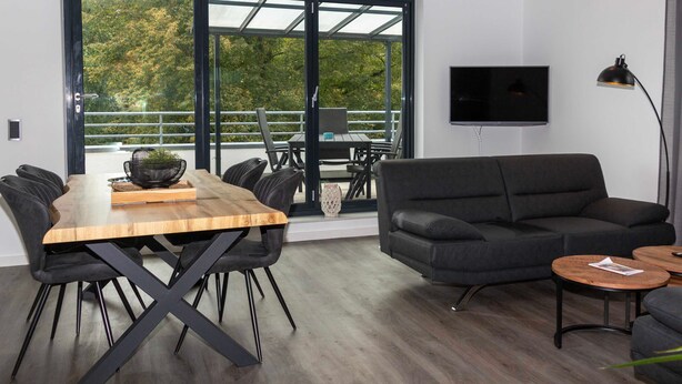 Una sala de estar moderna con una mesa de comedor de madera, sillas negras, un sofá gris y un televisor montado en la pared.