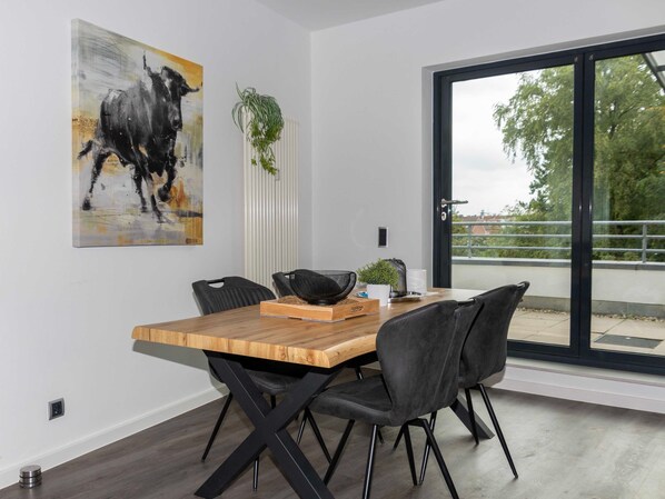 A modern dining area with a wooden table, black chairs, a black and white abstract painting, a potted plant, and a view through a large window.