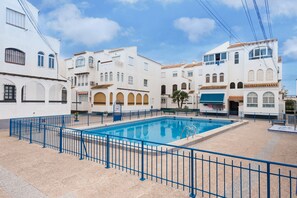 A swimming pool surrounded by white buildings with arched windows and balconies, a blue fence, and a clear sky.