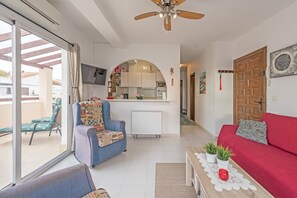 A living room with a red sofa, a blue armchair, a coffee table with plants, a ceiling fan, and a sliding glass door leading to a balcony.