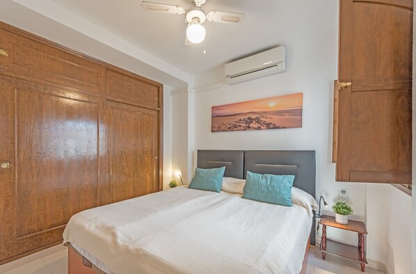 A bedroom with a large wooden wardrobe, a bed with a grey headboard, a small bedside table with a plant, and a wall-mounted air conditioner.