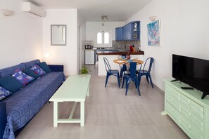 A modern living room with a sofa, a dining table, chairs, a TV, and a kitchen in the background.