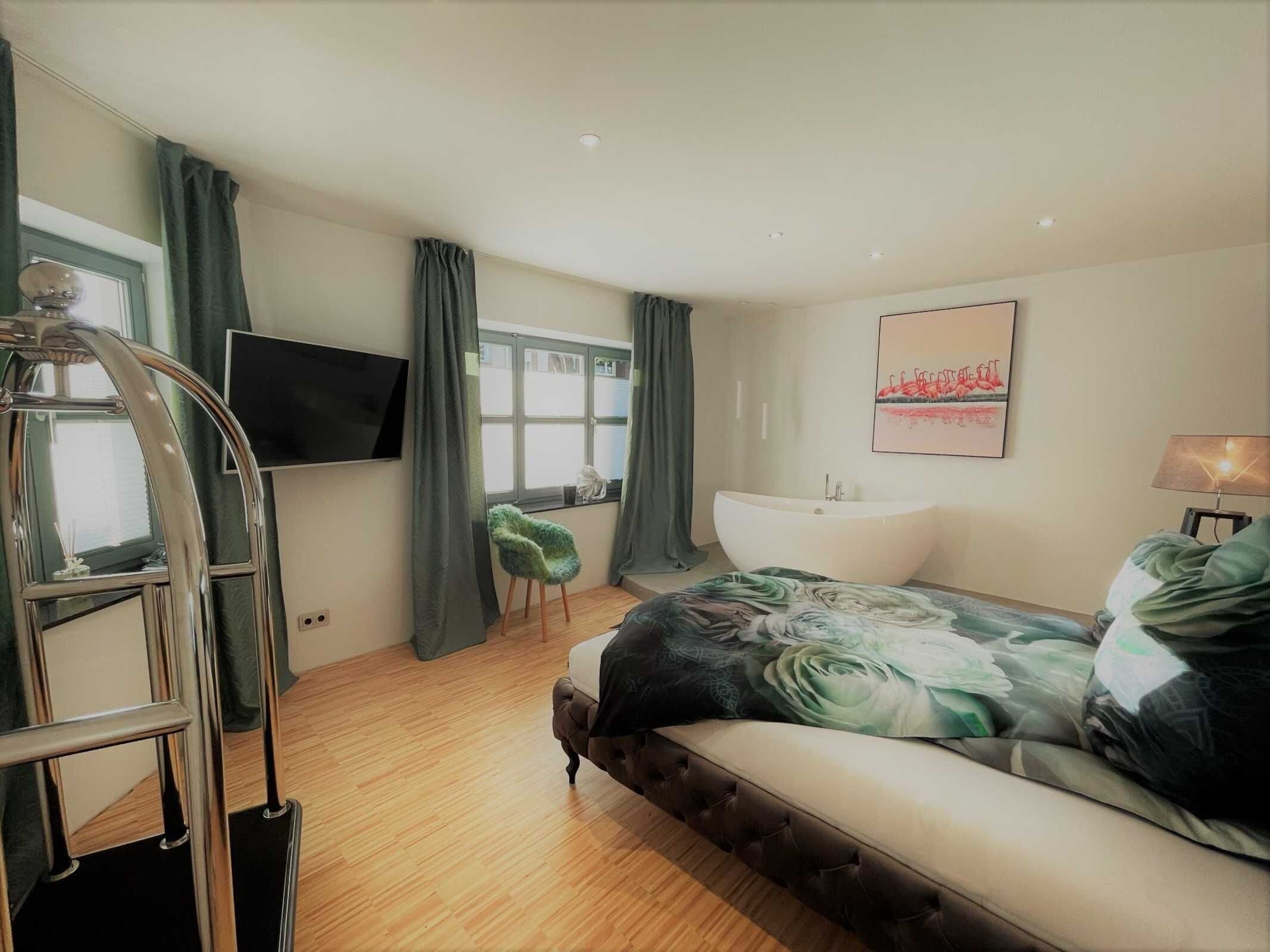 A bedroom with a bed, a TV, a window, a bathtub, and a chair.