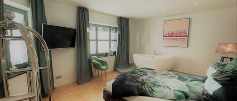 A bedroom with a bed, a TV, a window, a bathtub, and a chair.