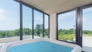 Un bain à remous sur un balcon doté de grandes fenêtres donnant sur un paysage verdoyant.