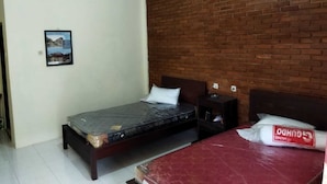 Un dormitorio con pared de ladrillo decorativa, una cama individual con colchón y almohadas, una mesita de noche con lámpara y una mesa roja con un bolso encima.