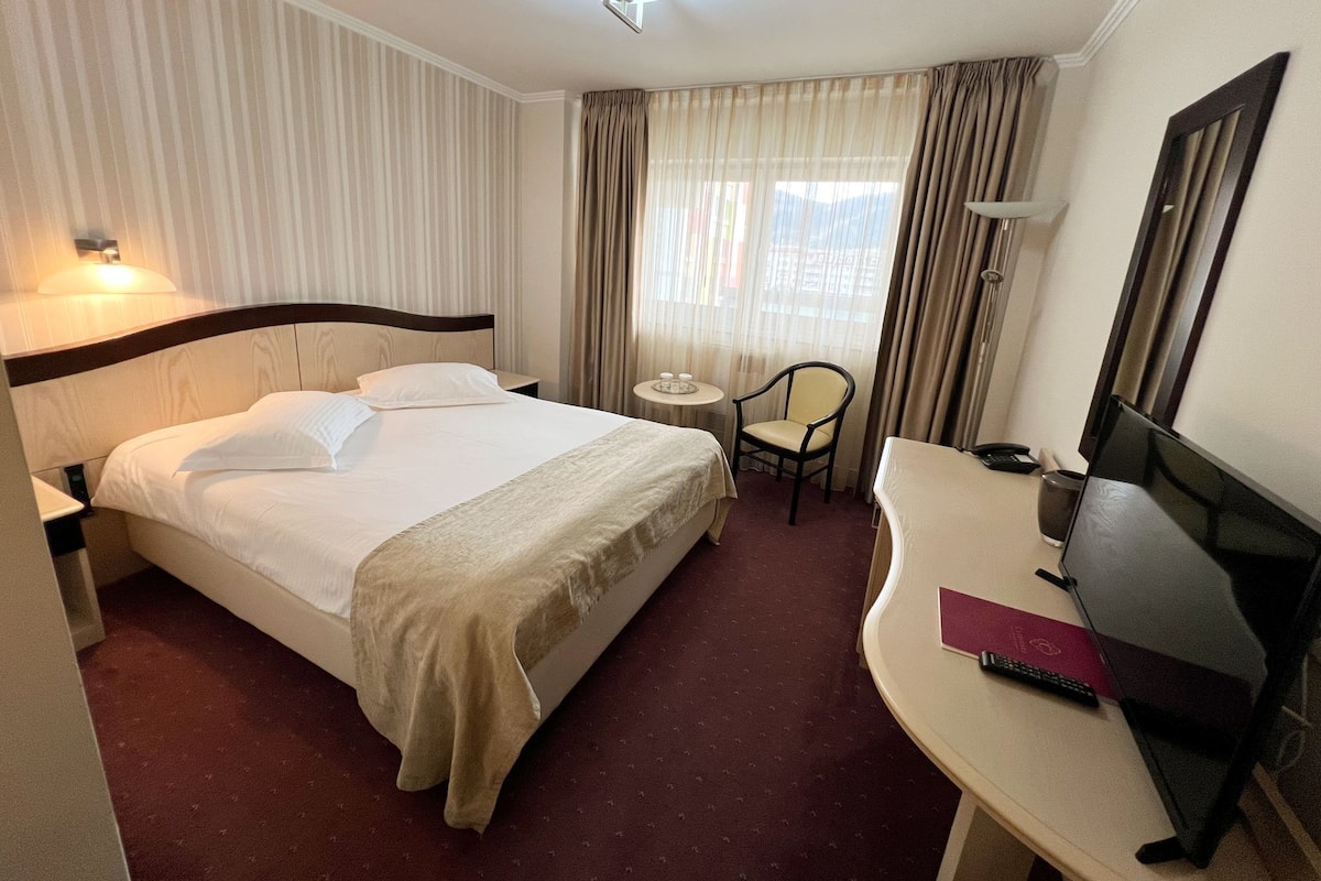 A hotel room with a bed, a desk with a TV, a chair, a small table with a lamp, and curtains.