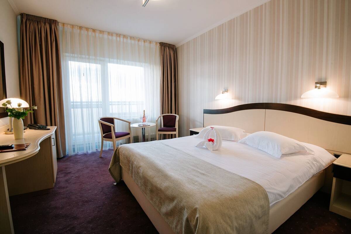 A hotel room with a large bed, a desk with a lamp, a chair, and a small table.