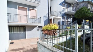 Un edificio residencial con entrada protegida por un portón, un pequeño jardín con plantas en macetas y un balcón visible.