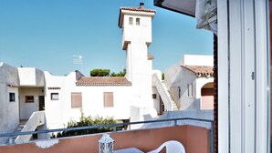 Un balcón con una mesa y dos sillas, con vista a un edificio blanco que tiene una torre y un tejado de tejas rojas.