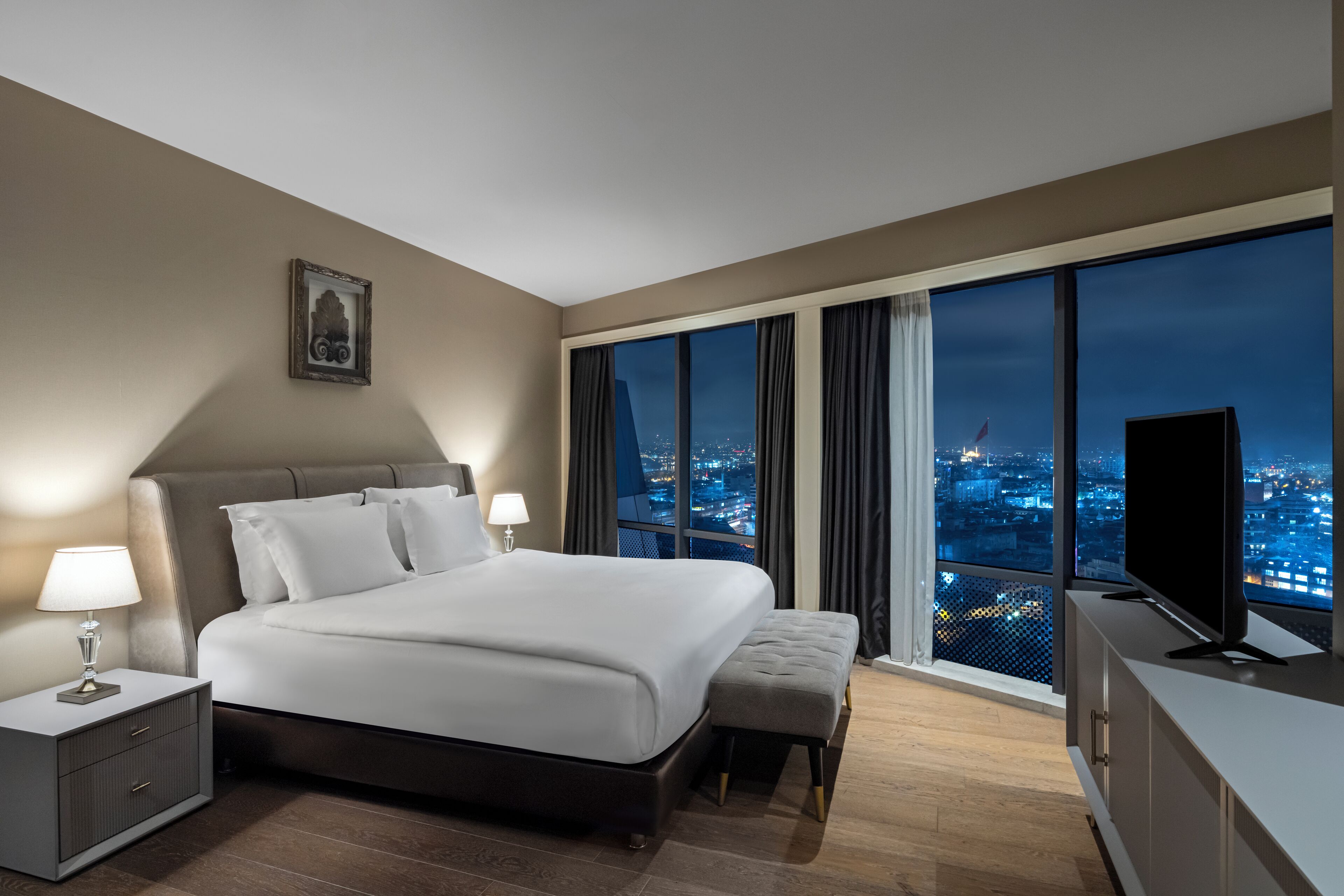 A modern hotel room with a large bed, a nightstand, a TV, and a view of a cityscape at night.