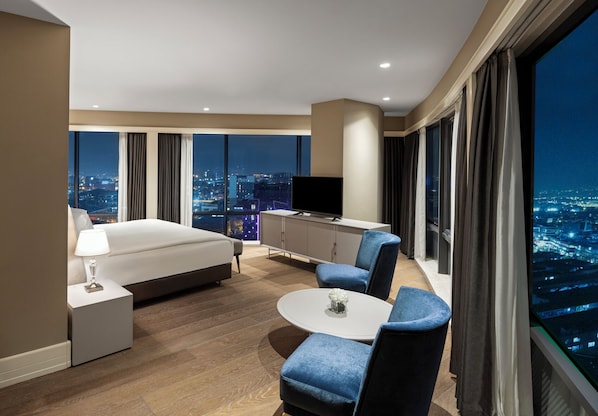 Une chambre d’hôtel moderne avec un grand lit, un bureau, une télévision et une vue sur la ville la nuit.
