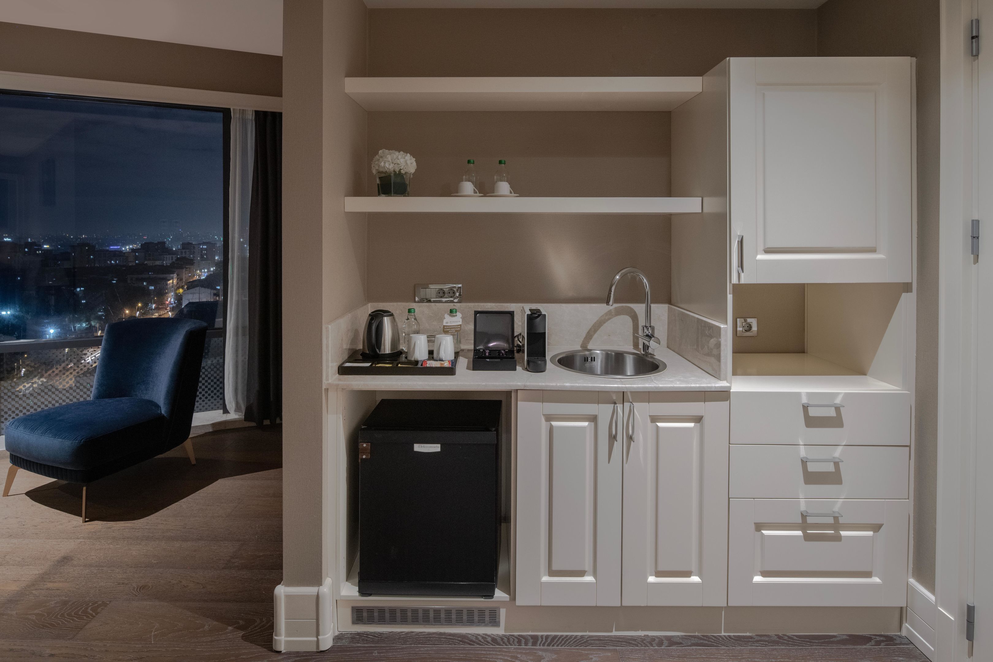 A compact kitchenette with a sink, countertop, and storage cabinets. A chair is positioned near the window, which offers a view of the cityscape at night.