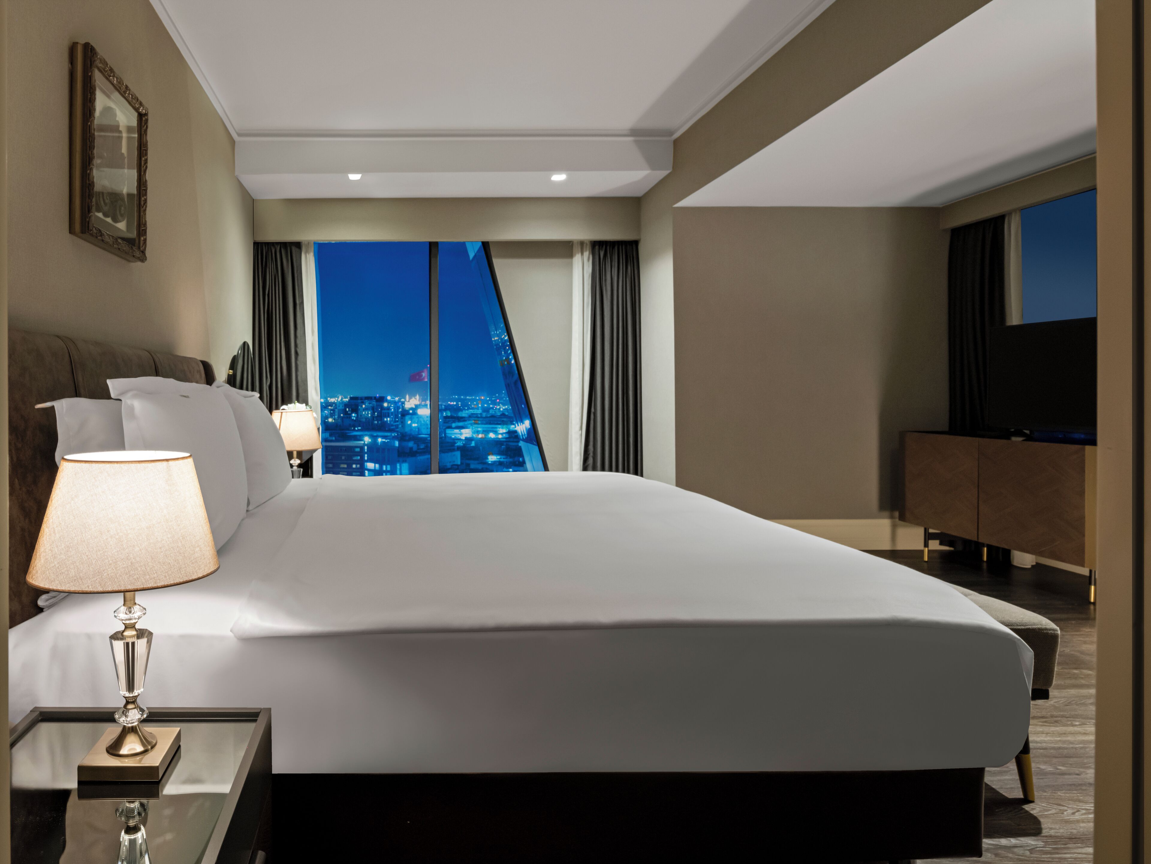 A modern hotel room with a large bed, a bedside table with a lamp, a TV on a cabinet, and a city view through the window.