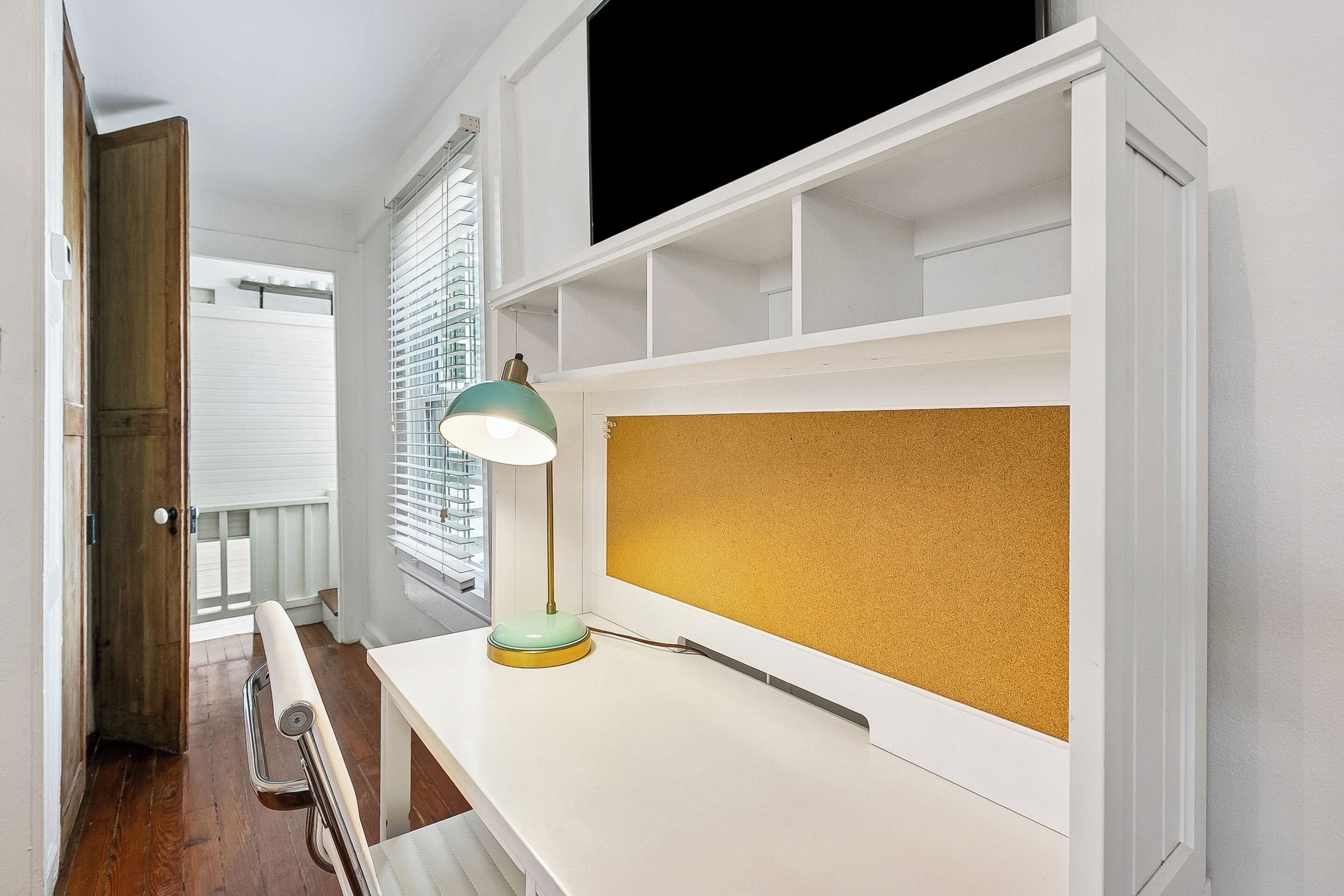 Une pièce avec un bureau blanc, un tableau d’affichage en liège, une lampe de bureau verte et une fenêtre avec des stores.