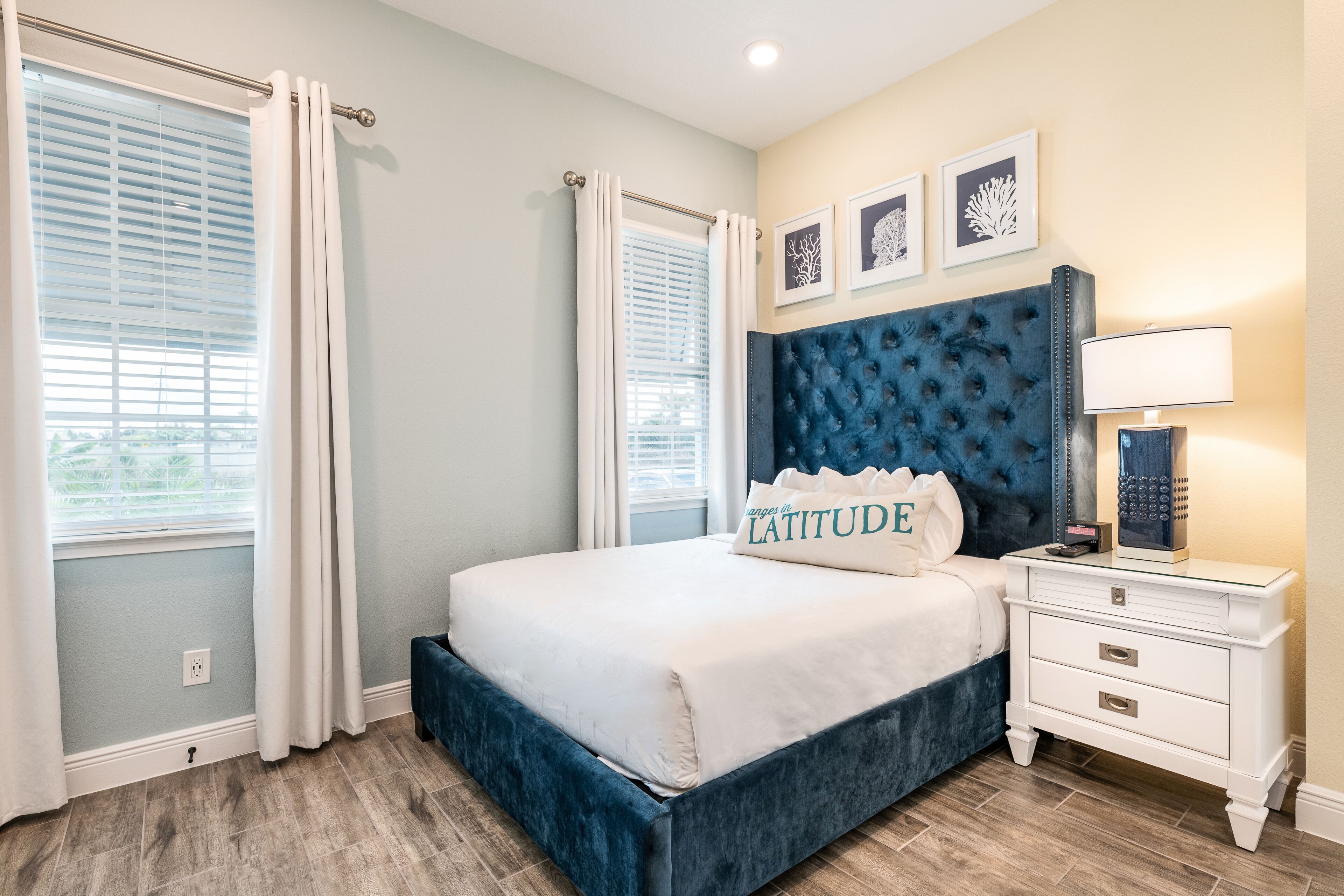 A bedroom with a bed, white curtains, a nightstand, a lamp, and framed artwork on the wall.