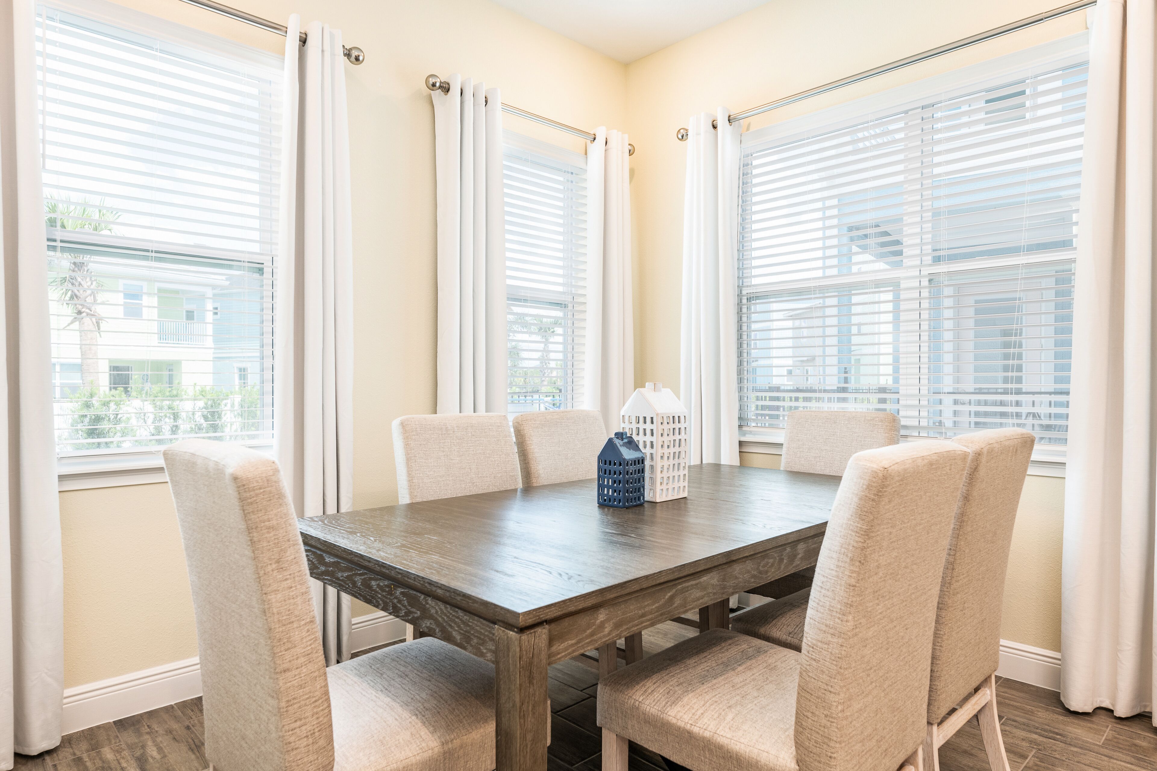 A dining area with a wooden table and six chairs, large windows with white curtains, and a view of buildings outside.