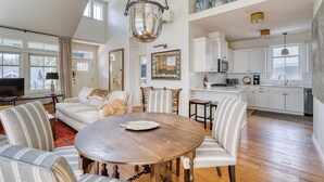 A spacious living room with a round wooden table, a sofa set, and a dining area with striped chairs. A kitchen with white cabinets and a large window is visible in the background. - Art Collector's Sophisticated NW Crossing Home (Bend)