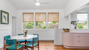 A room with a round table, teal chairs, a pink cabinet, and large windows with woven blinds.