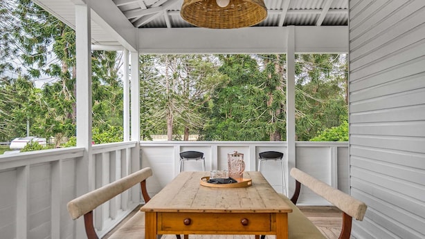 A wooden table with chairs and a wicker hanging light fixture in a covered porch with white railings and a view of trees.