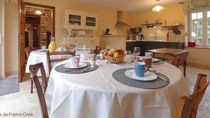 A dining area with a round table set for four, surrounded by wooden chairs. There is a kitchen in the background with modern appliances and a window with a view of greenery.