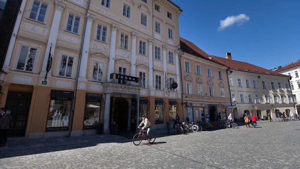 En historisk byggnad med flera butikslokaler, inklusive en väl synlig skylt med texten ”IKONA”, och en kullerstensgata där cyklar är uppställda längs trottoaren.