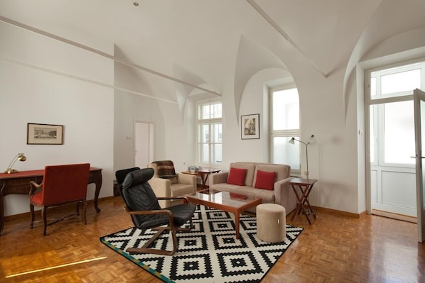 A spacious living room with a wooden floor, a black and white patterned rug, a red chair, a black leather sofa, a beige sofa with red cushions, a small round ottoman, and a desk with a lamp.
